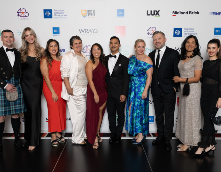 Group of ten people in formal evening attire posing together on a red-carpet style event backdrop featuring sponsor logos