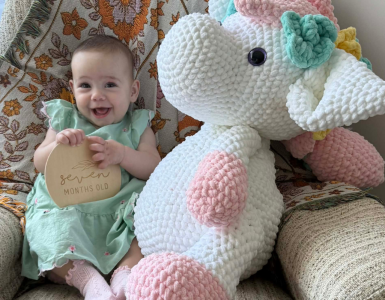 Baby sitting next to a unicorn soft toy.
