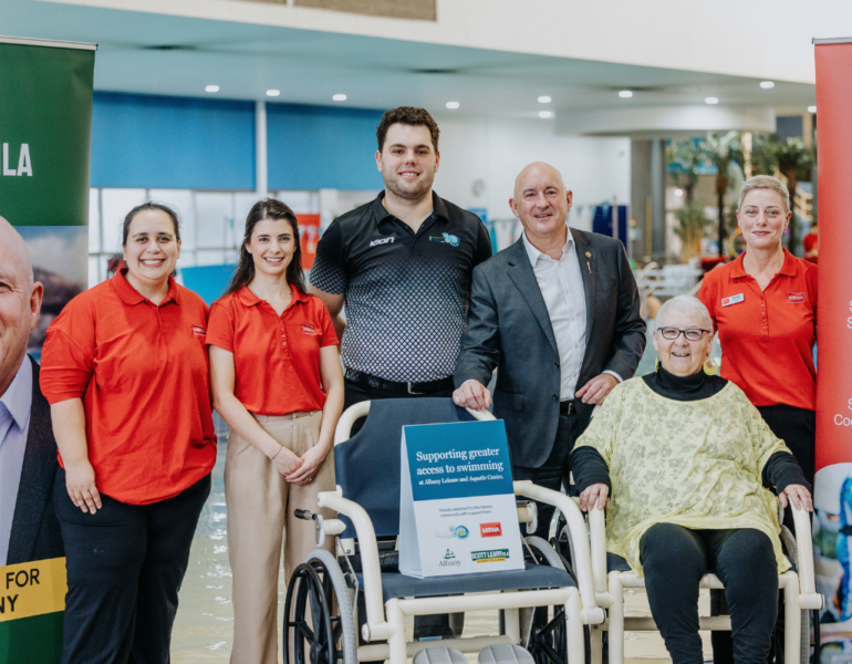 People at a pool standing next to a water wheelchair