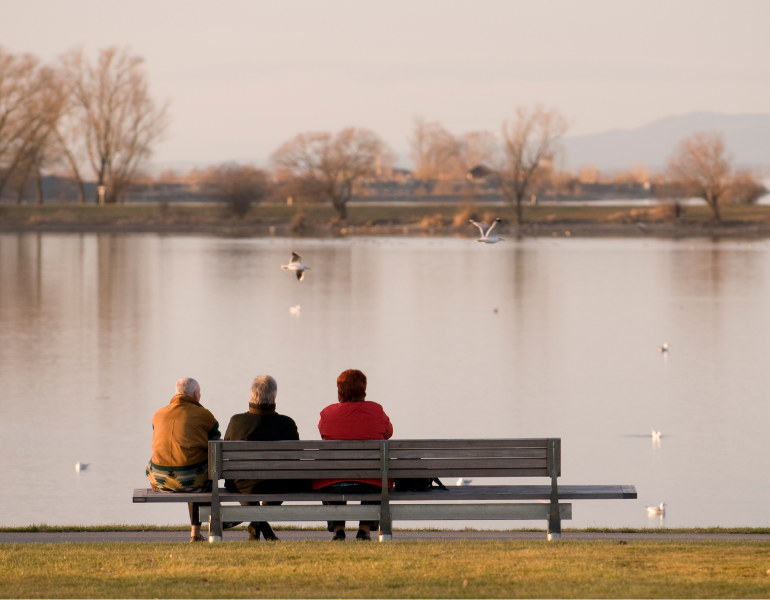 thee people sitting on a bench near a river or lake