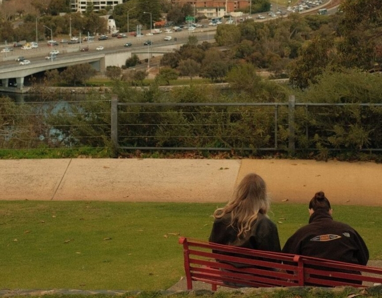 Two people sitting on a park bench looking over the Perth skyline.