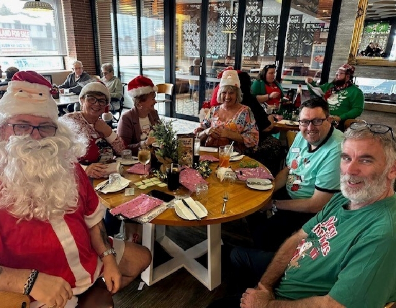 people sitting around a table, dresses in Christmas outfits, looking at the camera and smiling