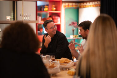 Man wearing glasses speaking to another man on left hand side at dining table.