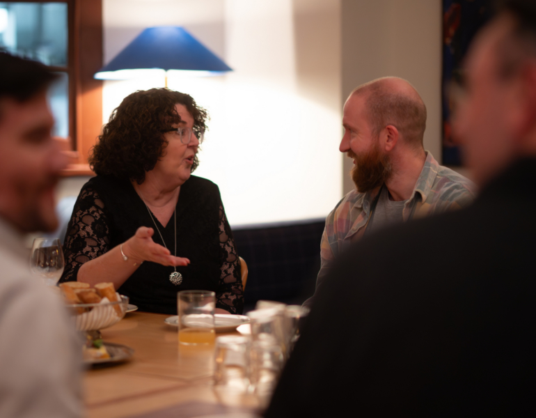 Lady on left speaking to man on the right at a dining table.