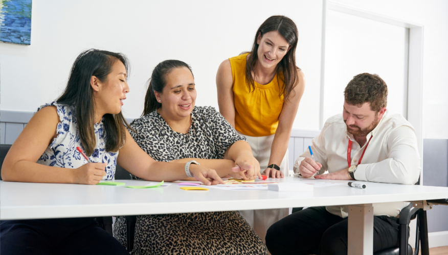 group of people sitting around a table and looking or pointing at something on the table