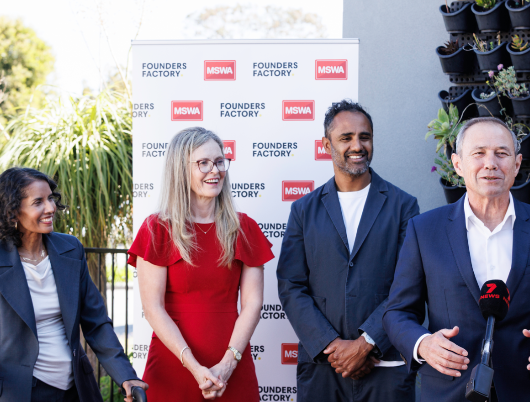 Nicolette, Melanie, Rohan and Roger Cook stand in front of a media wall speaking into a microphone.